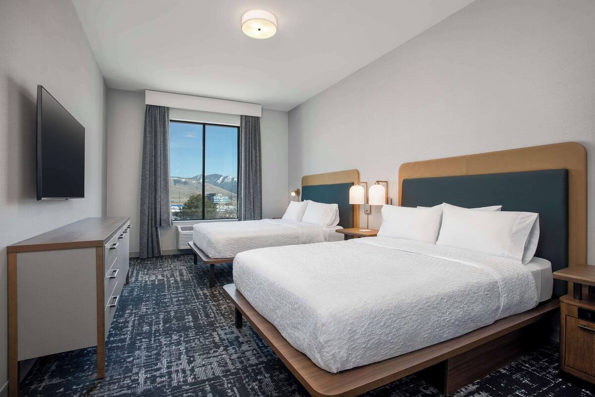 Habitación de hotel con dos camas, un escritorio, un televisor y una ventana con vistas a las montañas.