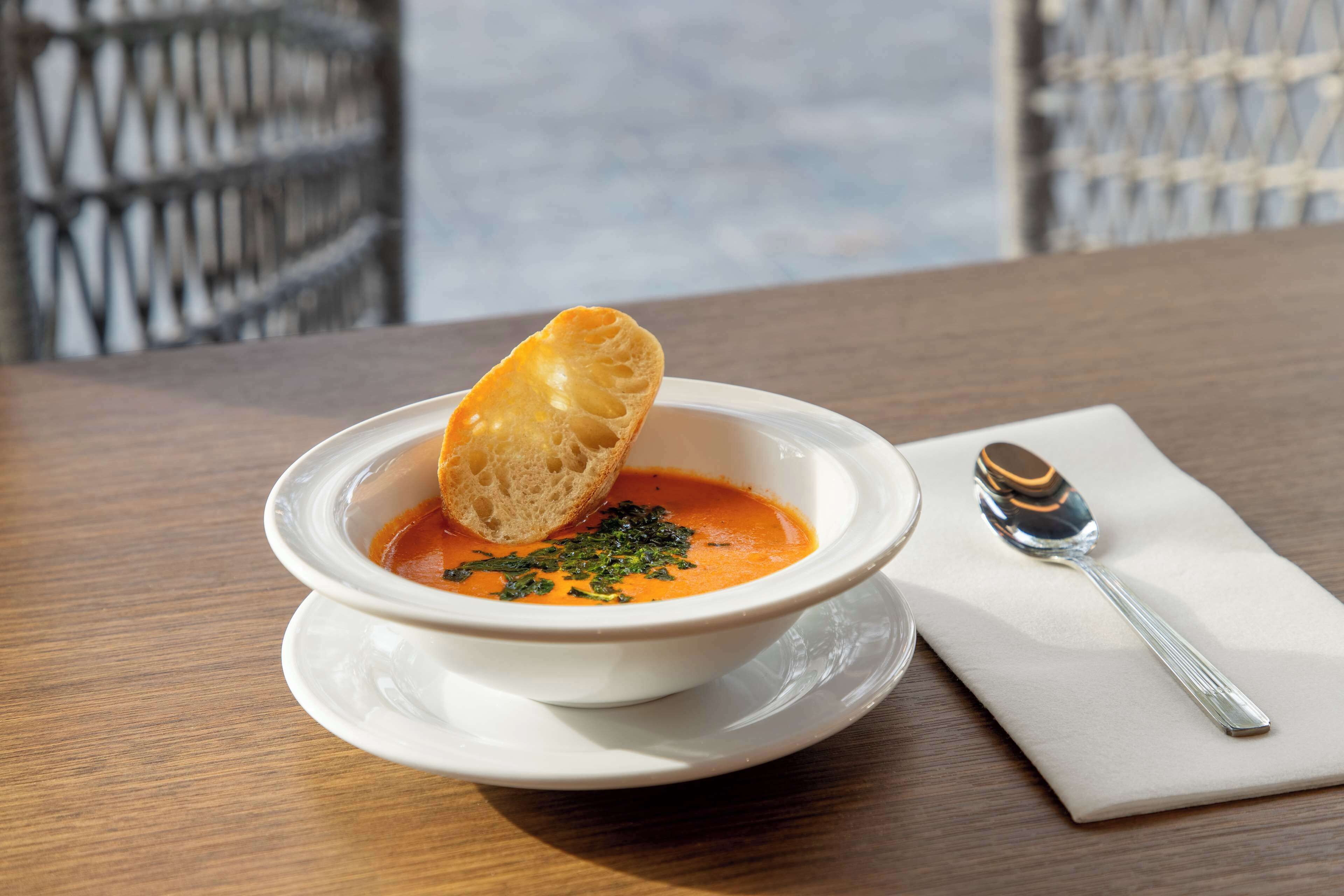 Un tazón de sopa con una rebanada de pan al lado, sobre un plato blanco en una mesa de madera.