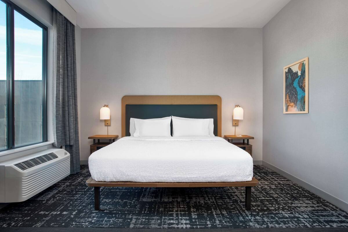 Habitación de hotel con una cama grande, dos lámparas de noche, una ventana con cortinas, un aire acondicionado instalado en la pared y un cuadro en la pared.