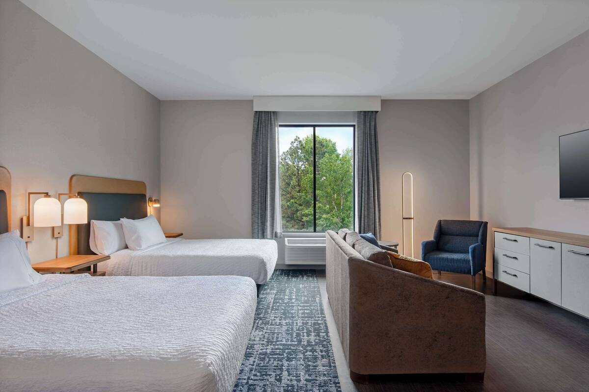 Habitación de hotel con dos camas, un sofá, un televisor y una ventana con vistas a árboles.