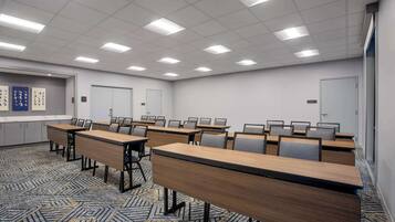 Un salón de conferencias con varias mesas y sillas, piso alfombrado e iluminación fluorescente en el techo.