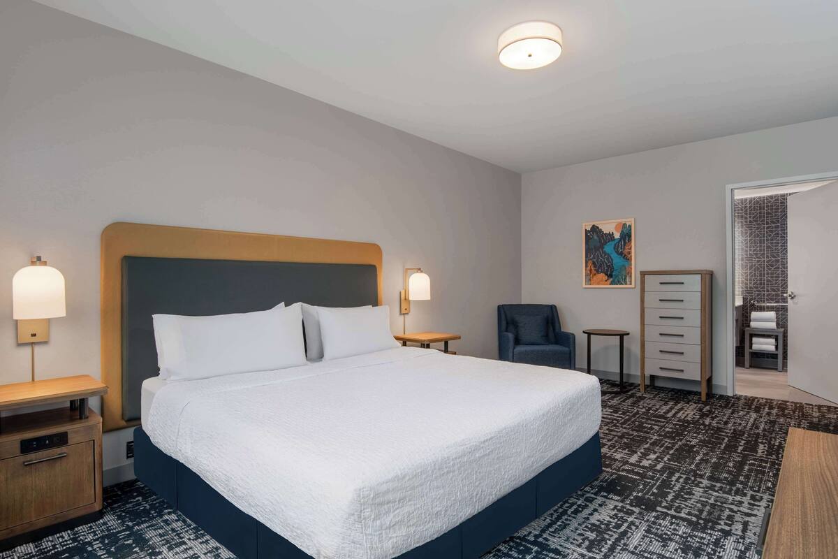 Una habitación de hotel con una cama grande, mesitas de noche, una silla, un aparador y un cuadro en la pared.