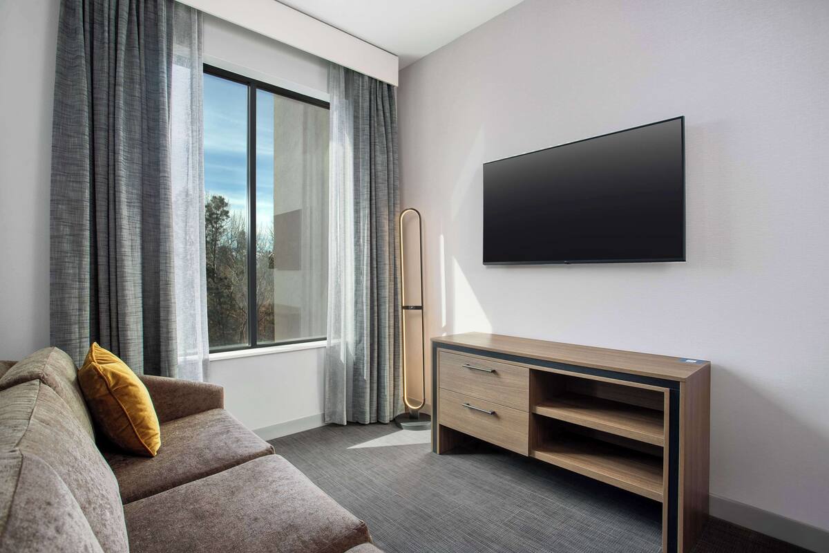 Habitación de hotel moderna con un gran ventanal, un televisor de pantalla plana montado en la pared, una mesa consola de madera y un sofá con un cojín color mostaza.