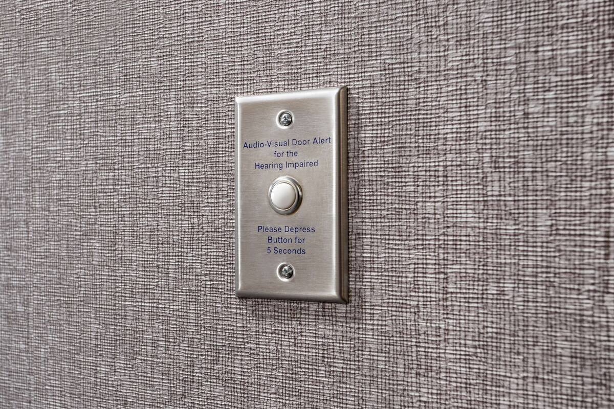 Interruptor metálico con la etiqueta 'Alerta de Puerta Audio-Visual para Personas con Discapacidad Auditiva', con instrucciones para presionar el botón durante 5 segundos.