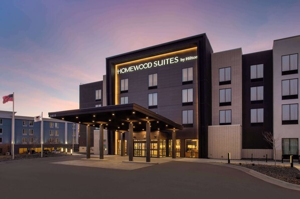 Een modern hotel met een bord waarop 'HOMEWOOD SUITES by Hilton' staat, en een overdekte ingang.