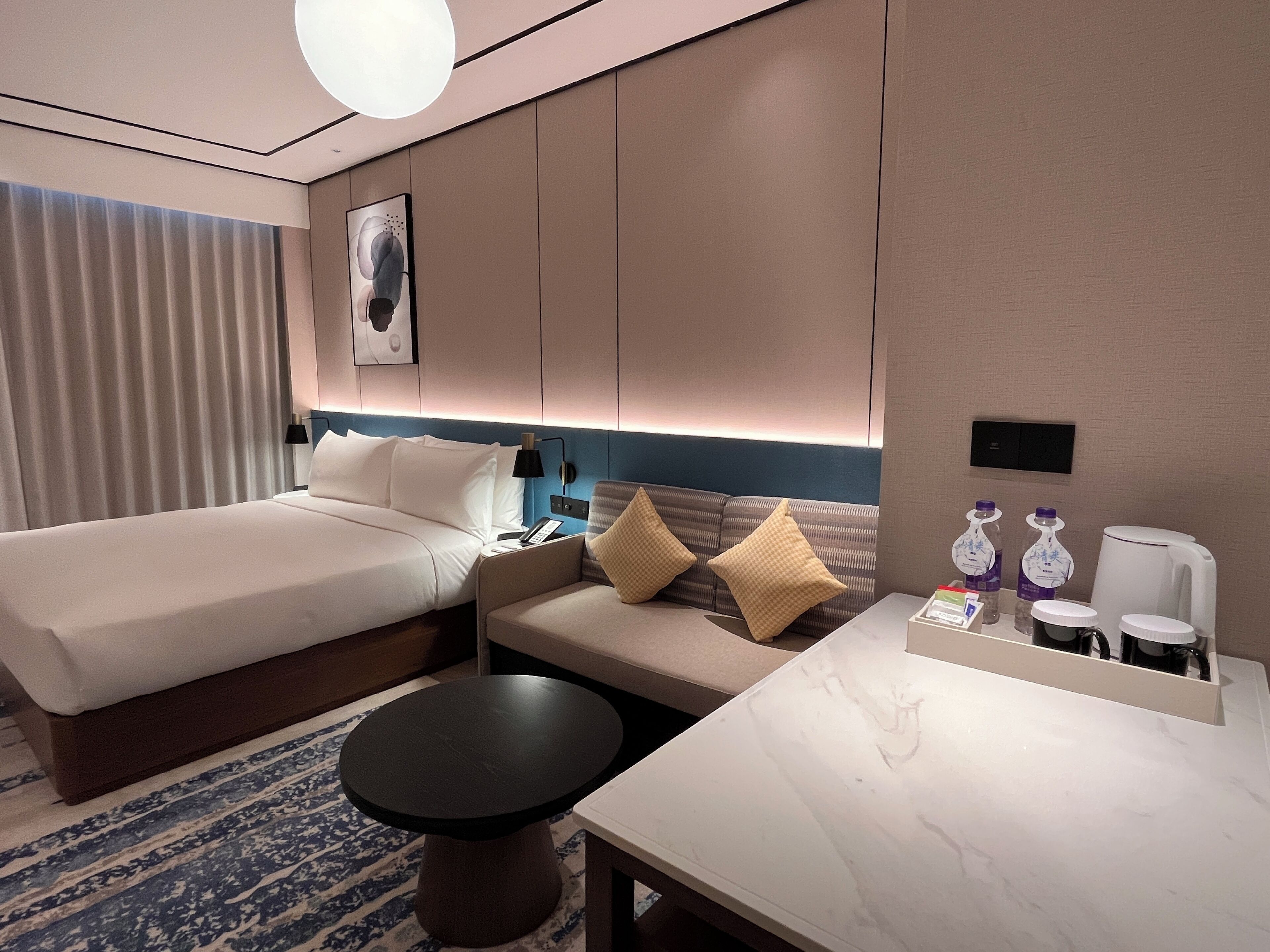 Habitación de hotel con cama, sofá, mesita, mesita de noche, lámpara y dispensador de agua.