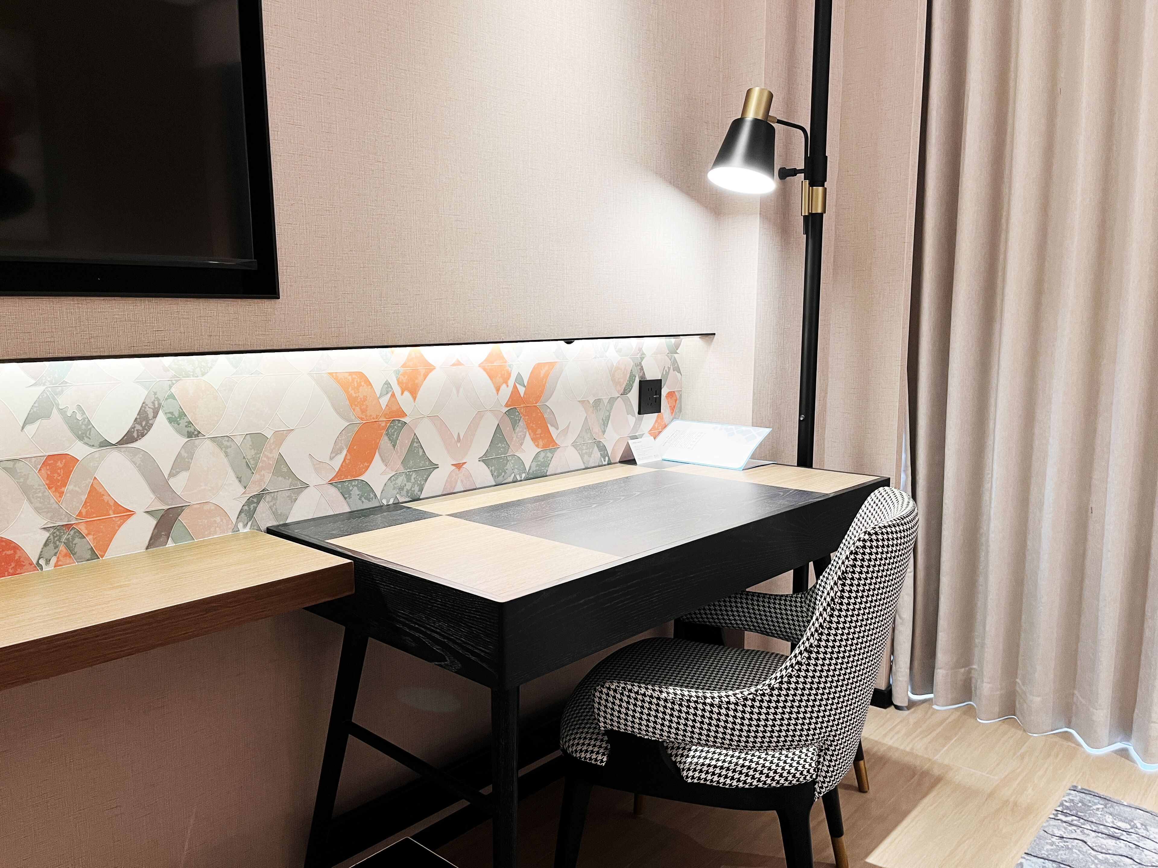 A modern room with a wooden desk, a black chair, a patterned wall, and a floor lamp.