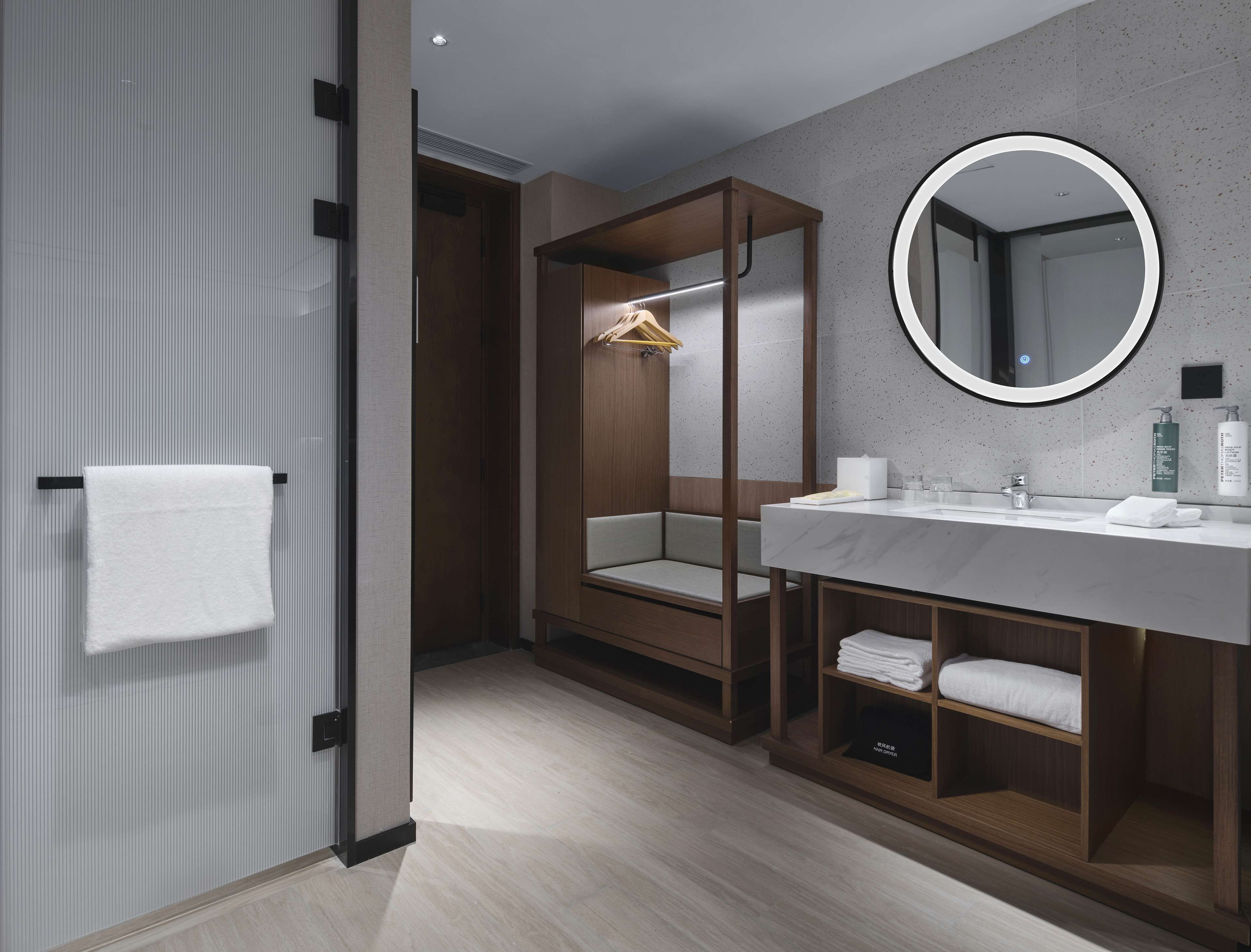 A modern bathroom with a wooden vanity, a round mirror, and a walk-in closet with a bench.