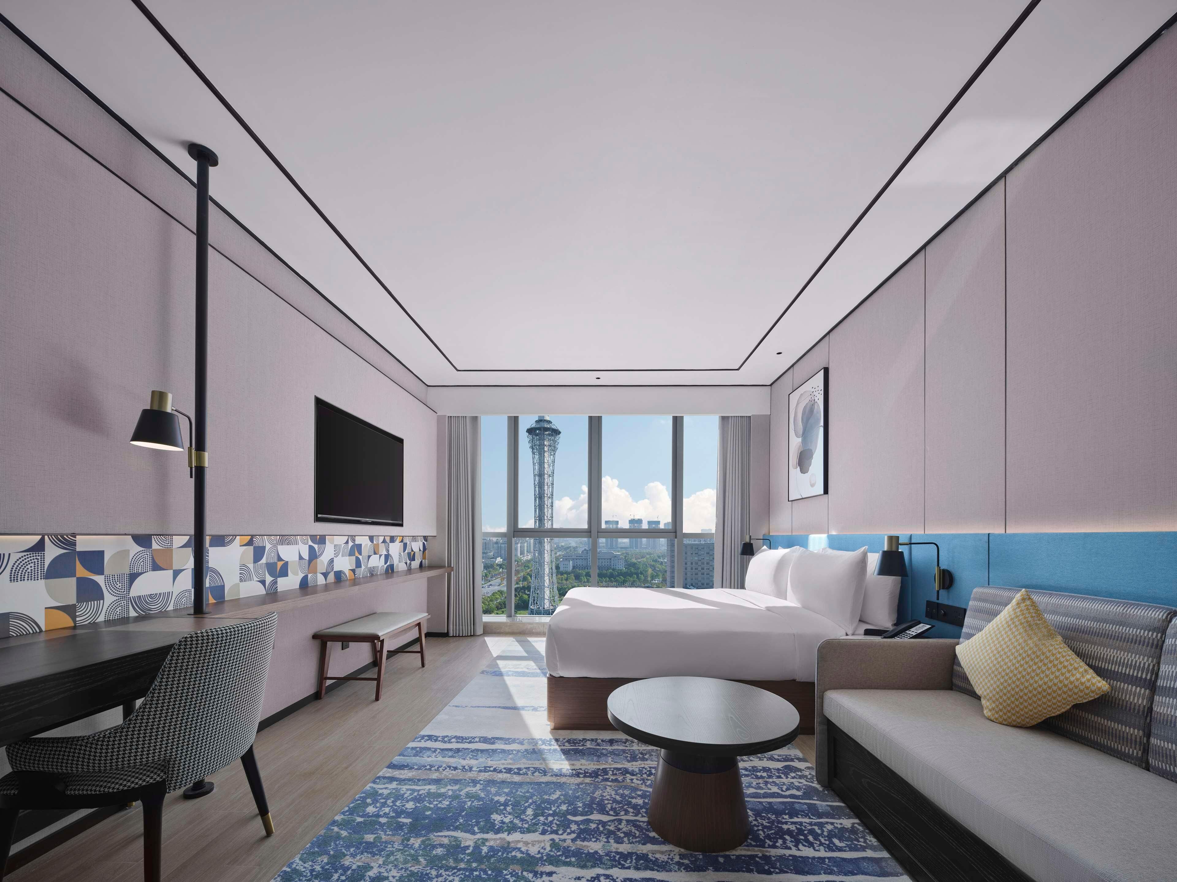 A modern hotel room with a large bed, a sofa, a dining table, and a TV mounted on the wall.