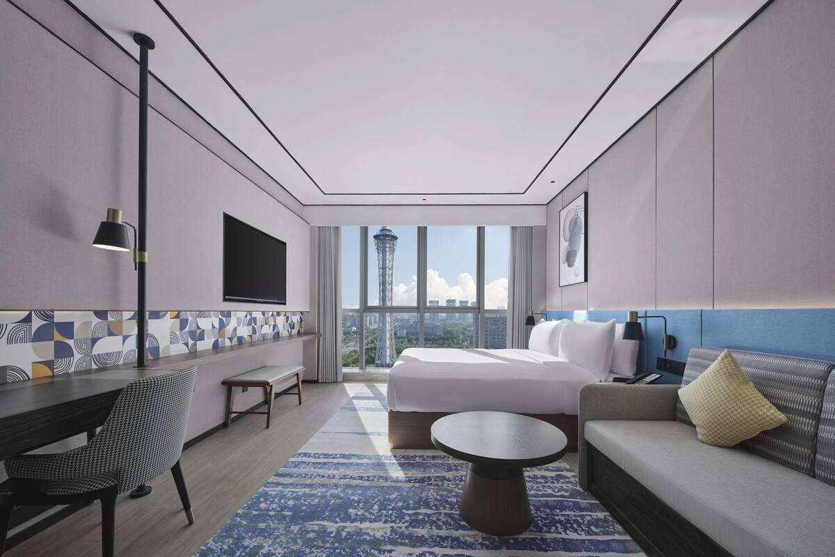 A modern hotel room with a large bed, a sofa, a coffee table, a desk, and a TV mounted on the wall.