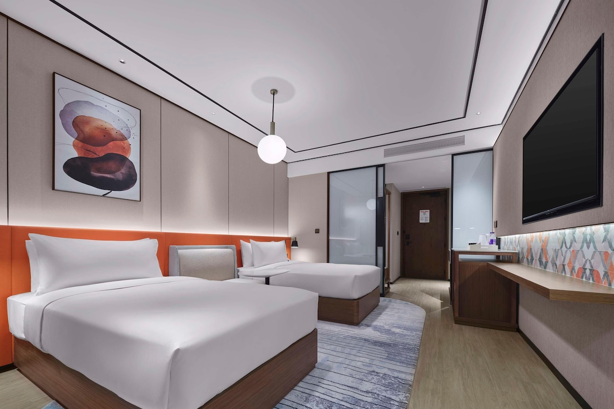 A hotel room with two beds, a large TV, a painting on the wall, and a desk with a computer.