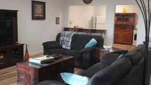 Ein Wohnzimmer mit einer Couch, einem Couchtisch und einem Fernseher. Im Hintergrund ist eine Küchenzeile mit Kühlschrank und Schränken zu sehen.