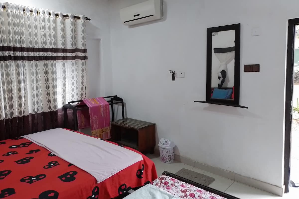 A bedroom with a bed, a chair, a small table, an air conditioner, a mirror, and a window with curtains.