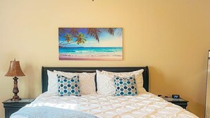 Una cama con colcha blanca, dos almohadas azules estampadas y un cuadro con una escena de playa encima.