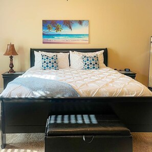 Un letto con una coperta bianca, due cuscini azzurri decorati e un quadro raffigurante una spiaggia sopra.