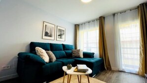 Ein Wohnzimmer mit einem dunkelgrünen Sofa, zwei gerahmten Bildern an der Wand, einem kleinen runden Tisch mit einem Buch und einer Pflanze sowie großen Fenstern mit Vorhängen.