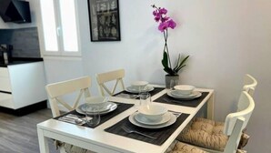 Ein Essbereich mit einem weißen Tisch, gedeckt für vier Personen, einschließlich Tellern, Gläsern und Besteck. Zwei Stühle mit gemusterten Kissen, ein Topfpflanze mit violetten Blüten und ein gerahmtes Bild an der Wand.