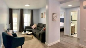 Ein modernes Wohnzimmer mit einer Couch, einem Couchtisch und einem Sessel. Eine Tür führt zu einem Schlafzimmer mit einem Bett.