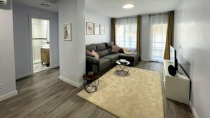 A modern living room with a grey sofa, a small round coffee table, and a beige rug. There is a white cabinet with a flat-screen TV on top. The room has wooden flooring and light grey walls. - Casa SoHo y Alex, Apartment 2A,- 2 bedroom (Estepona)