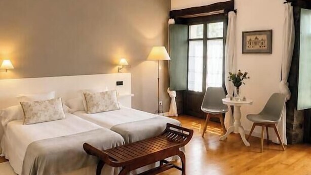 A hotel room with a bed, two bedside tables, a chair, a small table, and a window with curtains. - La Villa de Palacios (Voto)