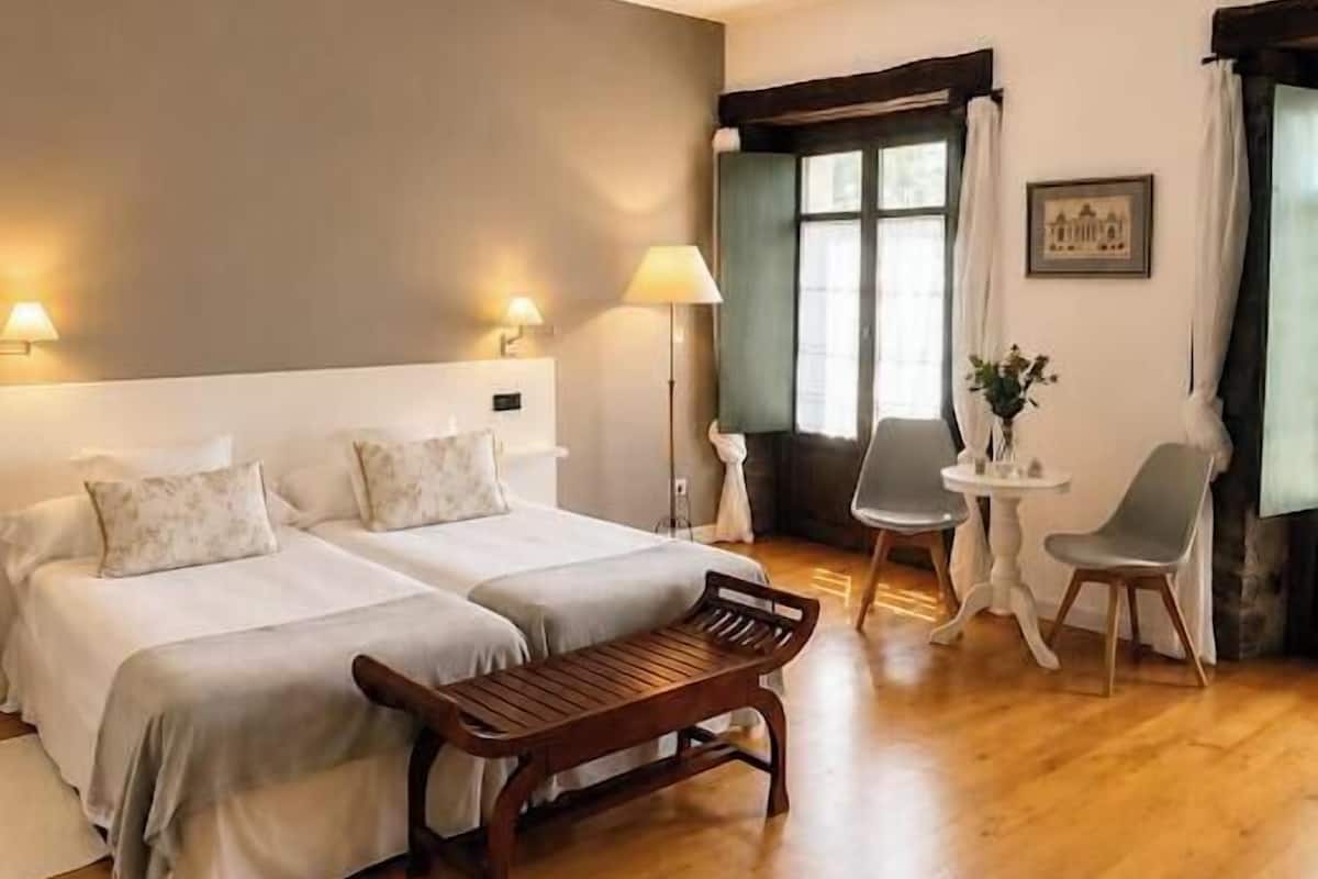A hotel room with a bed, two bedside tables, a chair, a small table, and a window with curtains.