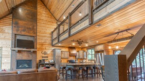 A spacious log cabin interior with a high ceiling, a central fireplace, a kitchen area, and a bar with stools. - 5Br cabin with private exterior bar and secret cinema from bunk room! (Broken Bow)