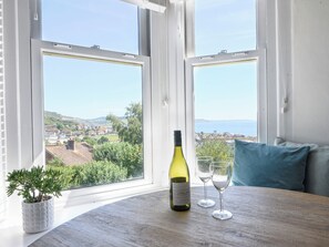 A room with a wooden table, a bottle of wine, and two glasses. A potted plant is on the windowsill, and a blue pillow is on the window seat. - Coastal View (Lyme Regis)