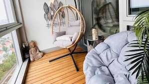 A balcony with a wooden floor, a grey cushioned chair, a wicker chair, and a Buddha statue. - Green High-end Apartment Close to Metro, Uno-city, E-parking, Pool, Gym, Sauna (Vienna)