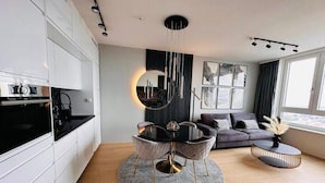 A modern kitchen with white cabinets, a black countertop, and a round dining table with chairs. There is a round mirror above the table and a sofa with cushions in the living area. - Green High-end Apartment Close to Metro, Uno-city, E-parking, Pool, Gym, Sauna (Vienna)