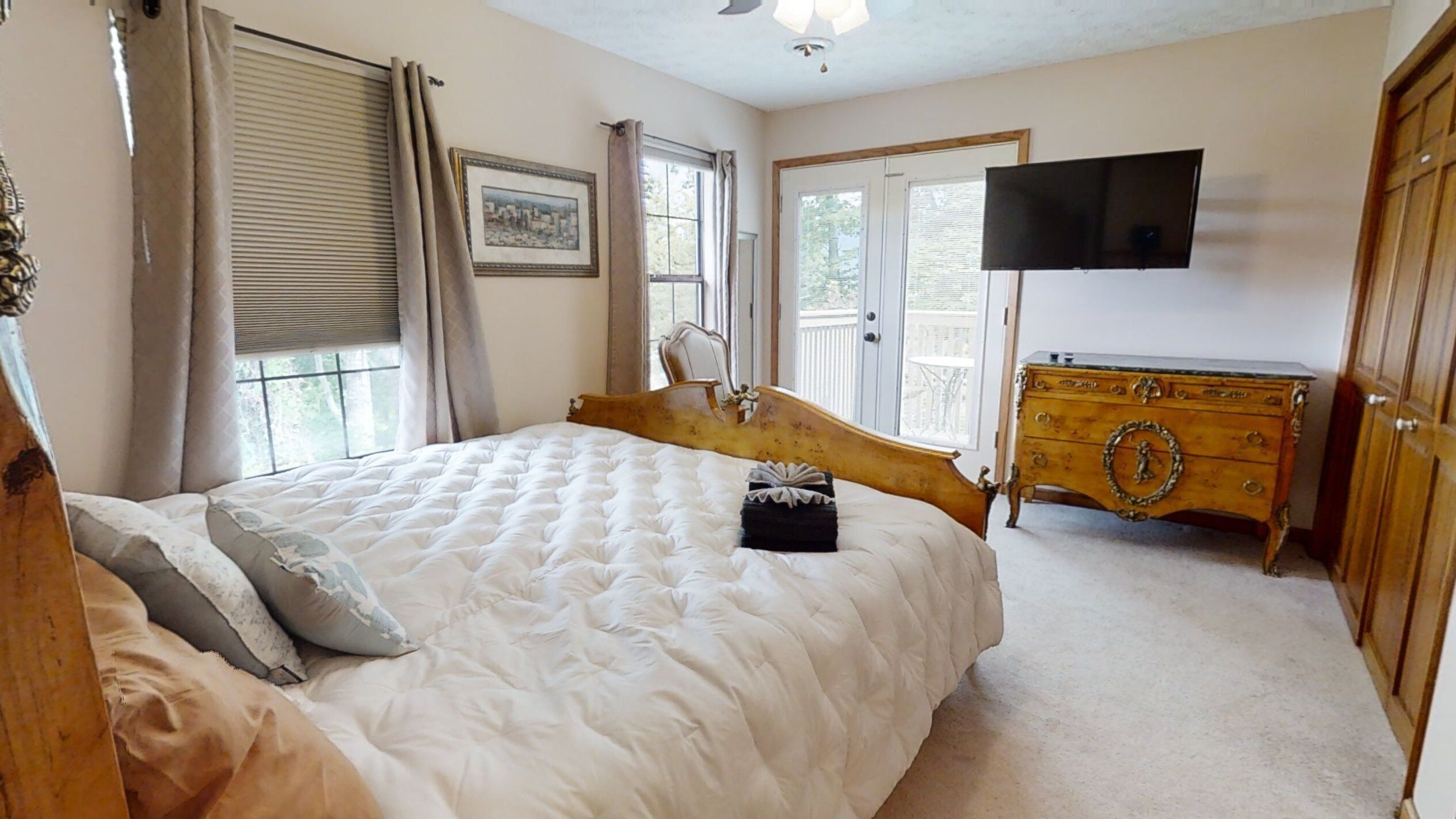 A bedroom with a large bed, a wooden dresser, a television mounted on the wall, and a window with curtains.
