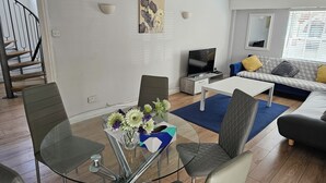 Een moderne woonkamer met een glazen eettafel, comfortabele zithoek, een flatscreen televisie en een trap.