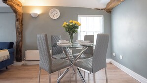 Een eetruimte met een glazen tafel, vier grijs gestoffeerde stoelen, een vaas met gele bloemen, een witte klok en een raam met jaloezieën.