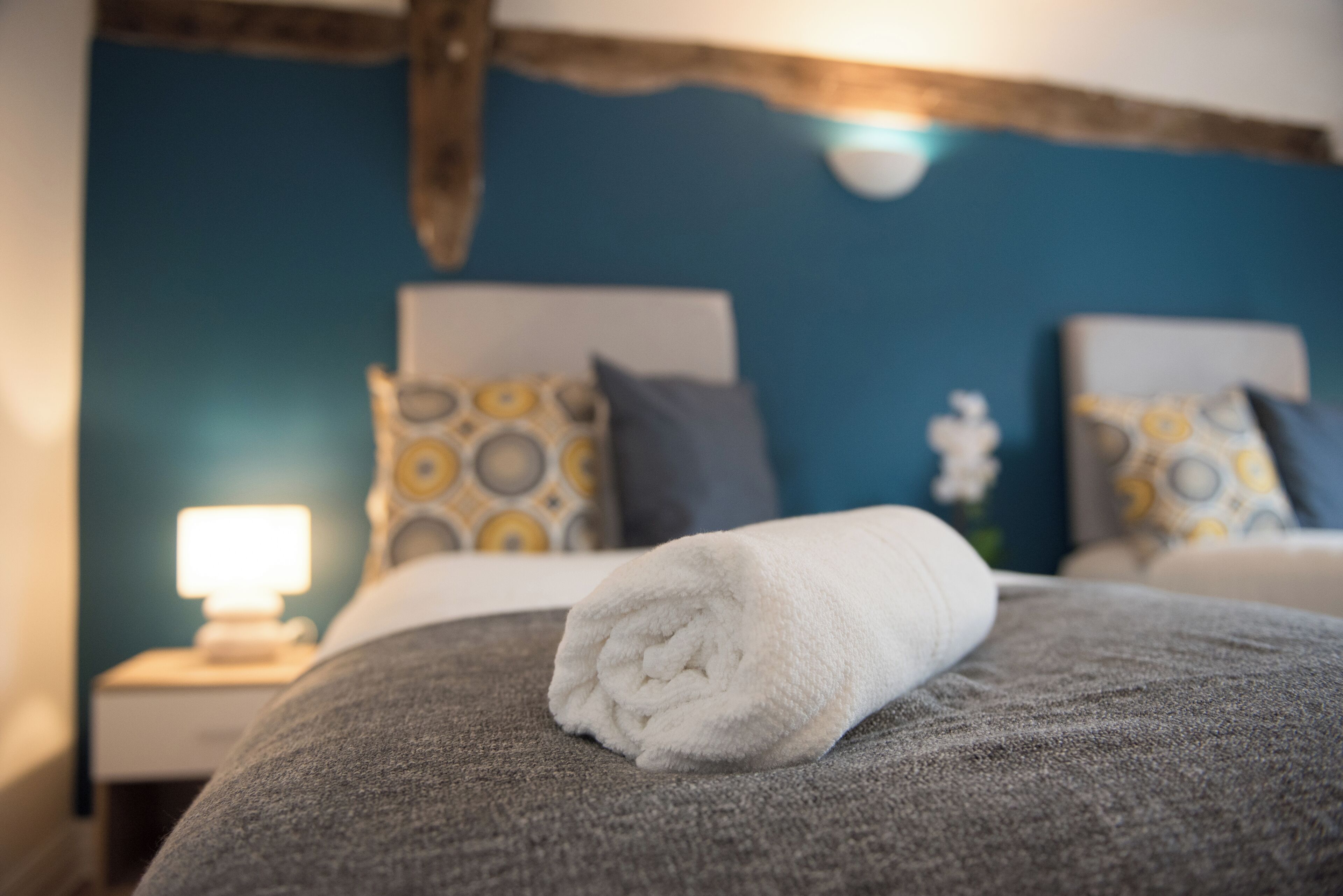 Un letto rifatto con cura, un asciugamano bianco arrotolato, una coperta grigia e un cuscino con un motivo giallo e bianco.
