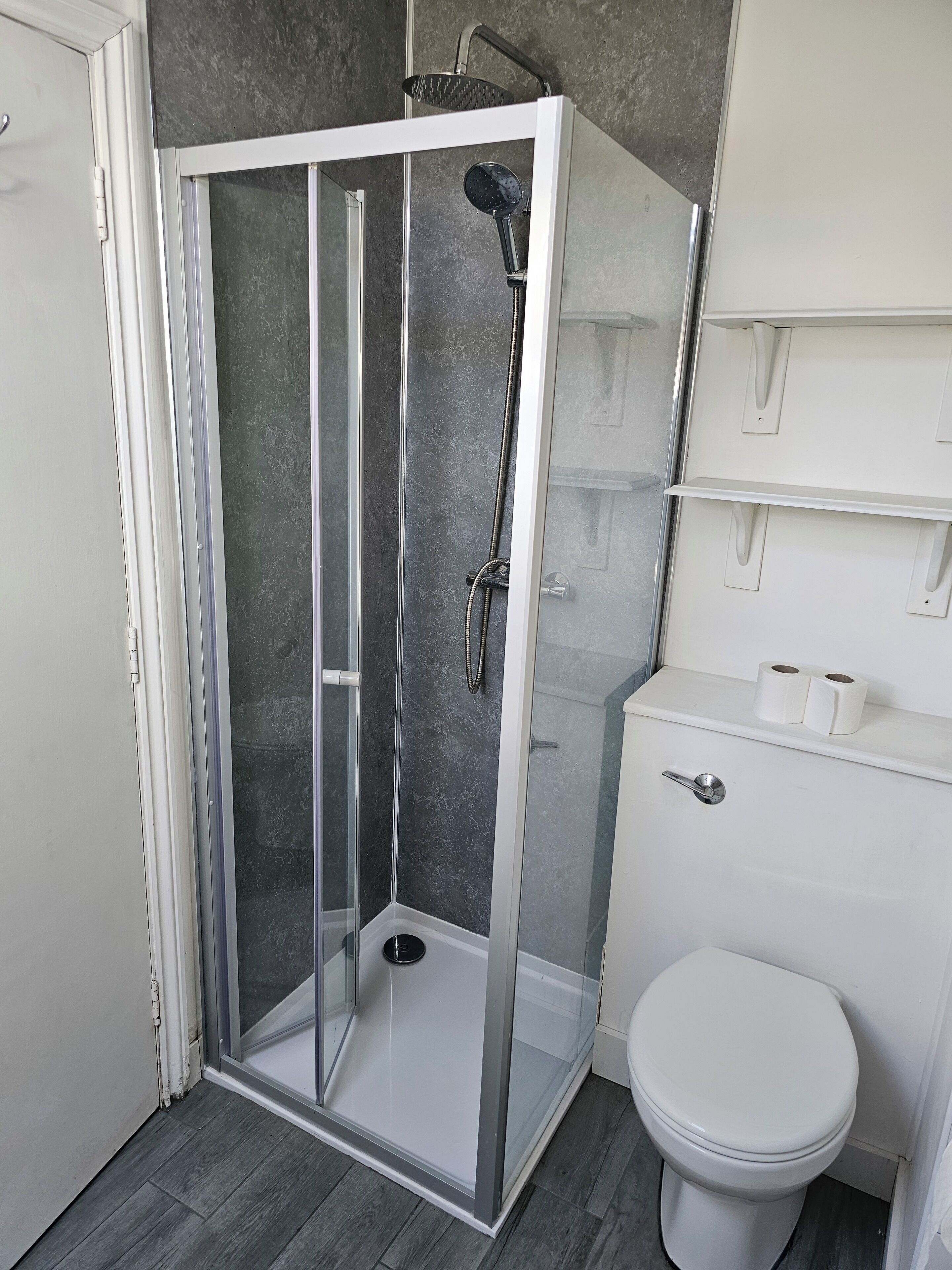 Un baño moderno con ducha de vidrio, inodoro y estante con papel higiénico.