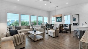 A spacious living room with a sofa, armchairs, a coffee table, and a dining area with chairs and a table. There is a ceiling fan, large windows, and artwork on the walls. - NEW!! Horizon! Private Beach ! Golf Cart ! Pool (Santa Rosa Beach)