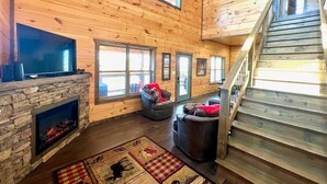 A cozy cabin interior with a stone fireplace, a flat-screen TV, and a staircase leading to an upper level. - Bearly Behaving (Sevierville)