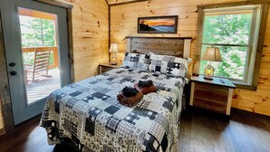 A wooden cabin bedroom with a bed, two bedside tables, a nightstand, a painting, and a window with a view of trees. - Bearly Behaving (Sevierville)
