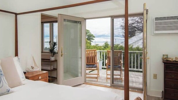 A bedroom with a bed, a nightstand, a dresser, and a sliding glass door leading to a balcony with chairs and a view of the outdoors. - Delightful & Modern seaside escape/Handicap Access (New London)