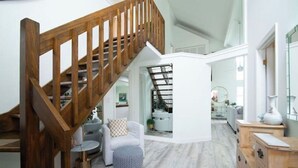 A spacious living area with a wooden staircase, a round coffee table, a white armchair, and a wooden cabinet. - Delightful & Modern seaside escape/Handicap Access (New London)