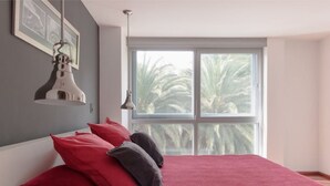 Une chambre avec une grande fenêtre, un lit avec des oreillers rouges et gris, et une suspension lumineuse.