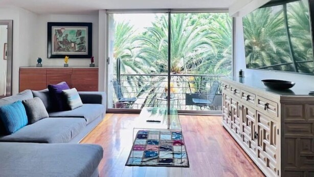 Un salon moderne avec un canapé gris, une table basse en verre et une vue sur des palmiers, visible à travers de grandes portes-fenêtres coulissantes.