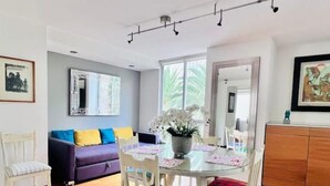 Un coin repas avec une table ronde, des chaises blanches et un vase de fleurs. On aperçoit un canapé avec des coussins colorés à l’arrière-plan.