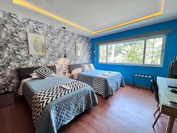 A bedroom with two beds, a desk, and a window with a view of greenery.