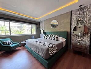 Une chambre à coucher moderne avec un grand lit, un fauteuil vert, une petite table et une grande fenêtre.
