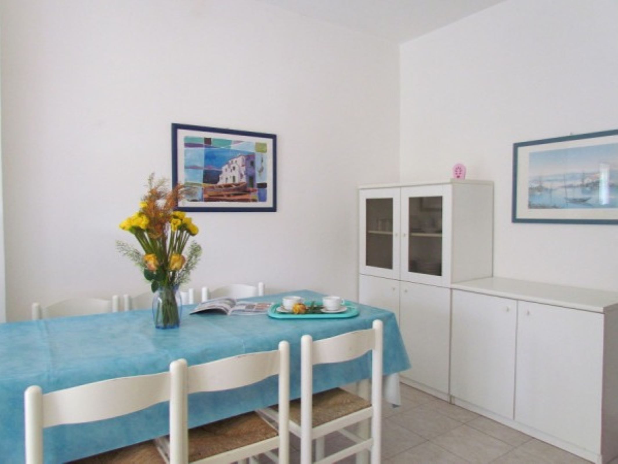 A dining area with a table covered by a blue tablecloth, white chairs, a vase with yellow flowers, and two framed pictures on the wall.