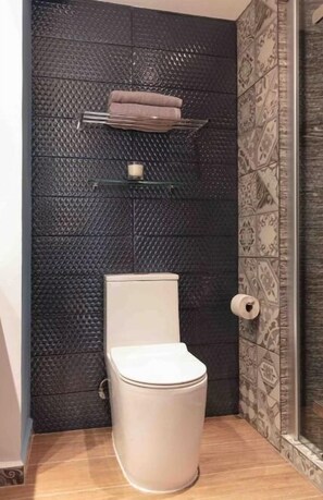 A modern bathroom with a textured dark wall, a white toilet, a glass shelf with toiletries, and patterned tiles on the floor and wall.