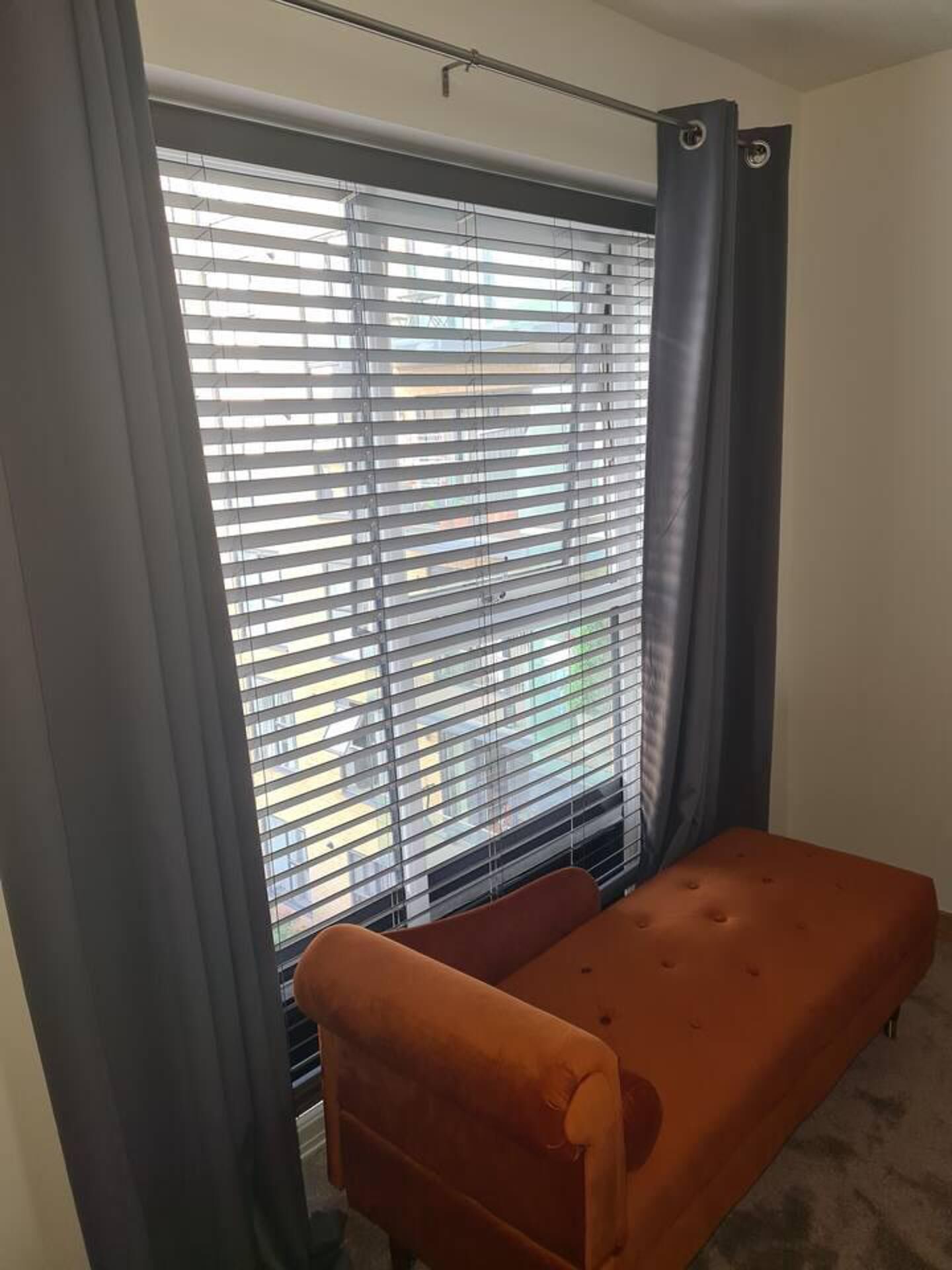 Una habitación con ventana cubierta por persianas, una cortina gris y una sola silla naranja.