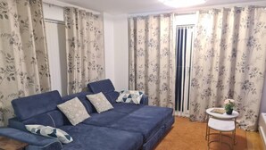 A cozy living room with a blue sofa, floral curtains, a small round table with a plant, and a window with blinds.