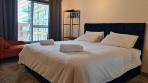 A hotel room with a large bed, a chair, a window with curtains, and a metal shelf.
