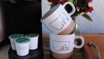Una cafetera Keurig, una taza con diseño de montaña y una taza de café sobre una mesa con un florero al fondo.